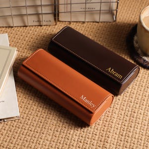 以下が含まれることがあります： 白ステッチが施された、茶色とタン色の2つのパーソナライズされた革製ケース。茶色のケースには金色の文字で「Abram」と刻まれ、タン色のケースには「Manley」と刻まれています。ケースは質感のある表面に置かれています。