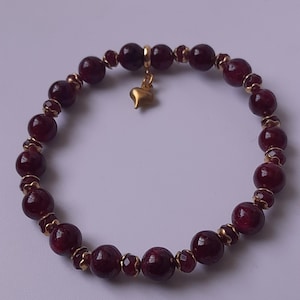 Peut inclure: Un bracelet de perles composé de perles sphériques polies rouge foncé alternant avec de plus petites perles rouges à facettes et des entretoises en métal doré. Un petit cœur lisse doré est suspendu au sommet du bracelet élastique.