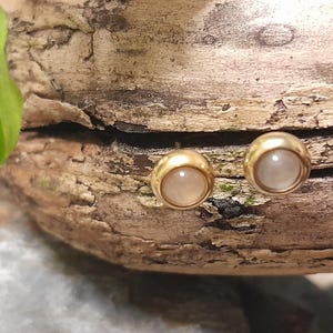 Peut inclure: Une paire de boucles d'oreilles à clous dorées, chacune ornée d'une pierre ronde et claire. Les boucles d'oreilles sont présentées sur un morceau de bois vieilli, mettant en valeur le design et la couleur des bijoux.
