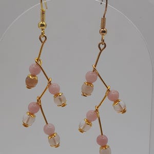 Peut inclure: Une paire de boucles d'oreilles pendantes dorées. Chaque boucle d'oreille présente un design de branche délicate avec des perles roses et transparentes. Les perles sont accentuées de capuchons dorés, créant un accessoire élégant et inspiré de la nature.