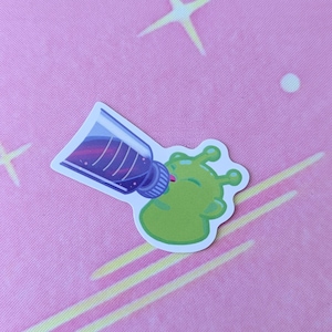 Op de afbeelding: Een sticker met een cartoon groene alien die uit een paarse fles drinkt. De alien heeft grote ogen en een glimlachende uitdrukking. De achtergrond is roze met witte sterren en gele en witte strepen.