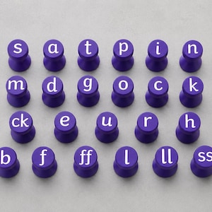 May include: A set of twenty-three purple circular tokens arranged in four rows, each featuring a white lowercase letter or letter combination. The letters displayed are s, a, t, p, i, n, m, d, g, o, c, k, ck, e, u, r, h, b, f, ff, l, ll, and ss.