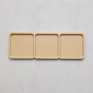 May include: A rectangular beige tray featuring three connected square compartments with rounded corners and raised edges. The tray is constructed as a single continuous piece with a smooth, matte finish and a uniform light tan colour throughout.