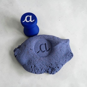 May include: A royal blue wooden stamp featuring the lowercase cursive letter a in white on its top surface, positioned next to a mound of purple kinetic sand that has been imprinted with the same lowercase cursive letter a design.