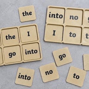 May include: Two tan rectangular trays with six square compartments each, containing tan square tiles printed with black text. The tiles feature the words to, the, I, no, go, and into. Several loose matching tiles are scattered around the trays.