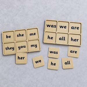 May include: Two light tan rectangular wooden trays with six square compartments each, containing black printed words. The words include be, she, me, they, you, my, her, was, we, are, he, and all. Several loose matching square wooden tiles are placed nearby.