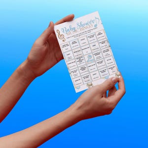 May include: A baby shower music bingo game card with a blue and gold design. The card features a grid of song titles and the text "Baby Shower Music Bingo". The card is held by two hands against a blue background.