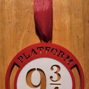 May include: A red and white circular ornament with the words "PLATFORM" and the numbers "9 3/4". The ornament is attached to a red ribbon. The design is reminiscent of a famous book series.