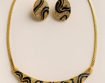 May include: A gold-toned necklace and matching earrings set. The necklace has a curved pendant with black enamel and crystal accents. The earrings are oval-shaped with a similar design. The set is on a light background.