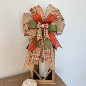 Könnte beinhalten: Eine dekorative Herbstschleife mit Burlap-, Karo- und einfarbigen Bändern in Orange, Grün und Braun. Die Schleife befindet sich auf einer Holzlaterne mit einer weißen Kerze im Inneren. Daneben liegt ein kleiner weißer Kürbis.