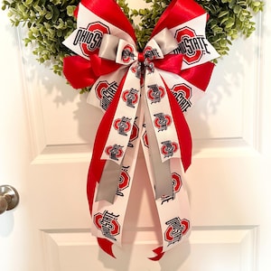 May include: A decorative wreath featuring a large bow with red, white, and gray ribbons. The bow is adorned with the text "OHIO STATE" and the Ohio State University logo. The wreath is made of green foliage and is hanging on a white door.