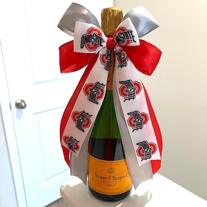 May include: A bottle of Veuve Clicquot champagne adorned with a decorative bow made of red, silver, and white ribbons. The white ribbon features the Ohio State logo and text printed in black and red, while the red and silver ribbons are solid.