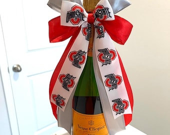 May include: A bottle of Veuve Clicquot champagne adorned with a decorative bow made of red, silver, and white ribbons. The white ribbon features the Ohio State logo and text printed in black and red, while the red and silver ribbons are solid.
