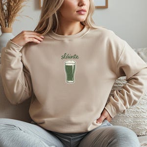 May include: Beige sweatshirt with the word "sl&aacute;inte" above a green pint of beer graphic. The graphic has a white foamy top. The sweatshirt is a neutral color and has long sleeves. The design is centered on the chest.