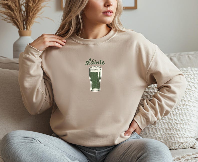 May include: Beige sweatshirt with the word "sl&aacute;inte" above a green pint of beer graphic. The graphic has a white foamy top. The sweatshirt is a neutral color and has long sleeves. The design is centered on the chest.