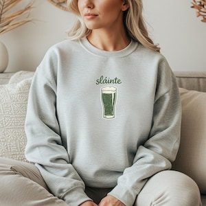 May include: A light gray sweatshirt with the word "sl&aacute;inte" above a green pint of beer. The beer has a white foamy head. The sweatshirt is made of a soft material.
