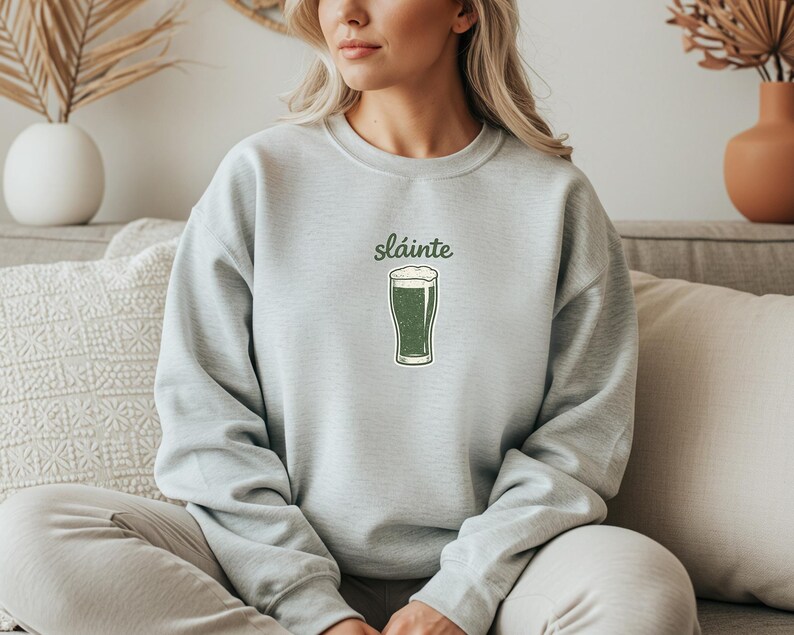 May include: A light gray sweatshirt with the word "sl&aacute;inte" above a green pint of beer. The beer has a white foamy head. The sweatshirt is made of a soft material.