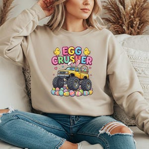 Op de afbeelding: Beige sweatshirt met een kleurrijke afbeelding met een gele monstertruck met konijnenoren, de tekst "EGG CRUSHER" en paaseieren. Het ontwerp bevat gele kuikens en harten.