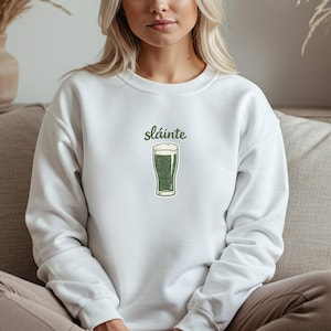 White-colored t-shirt with a small minimalist dark green line art drawing of a pint of stout beer and the handwritten-style Irish word "Sláinte" above it.