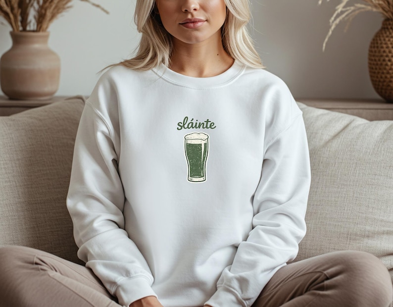 White-colored t-shirt with a small minimalist dark green line art drawing of a pint of stout beer and the handwritten-style Irish word "Sláinte" above it.