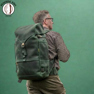 Può includere: Uno zaino in pelle verde scuro con patta superiore, cinghie regolabili e fibbie in metallo. La borsa presenta tasche laterali e una chiusura centrale a cinghia. Un logo mostra un sacco da boxe e il testo Punching Bags Heaven.