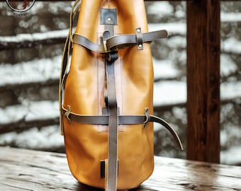 Könnte beinhalten: Ein hellbrauner Lederrucksack mit braunen Riemen und Schnallen. Die Tasche hat einen Kordelzugverschluss an der Oberseite und einen vertikalen Riemen in der Mitte. Das Logo "PUNCHING BAGS HEAVEN" ist oben links sichtbar.