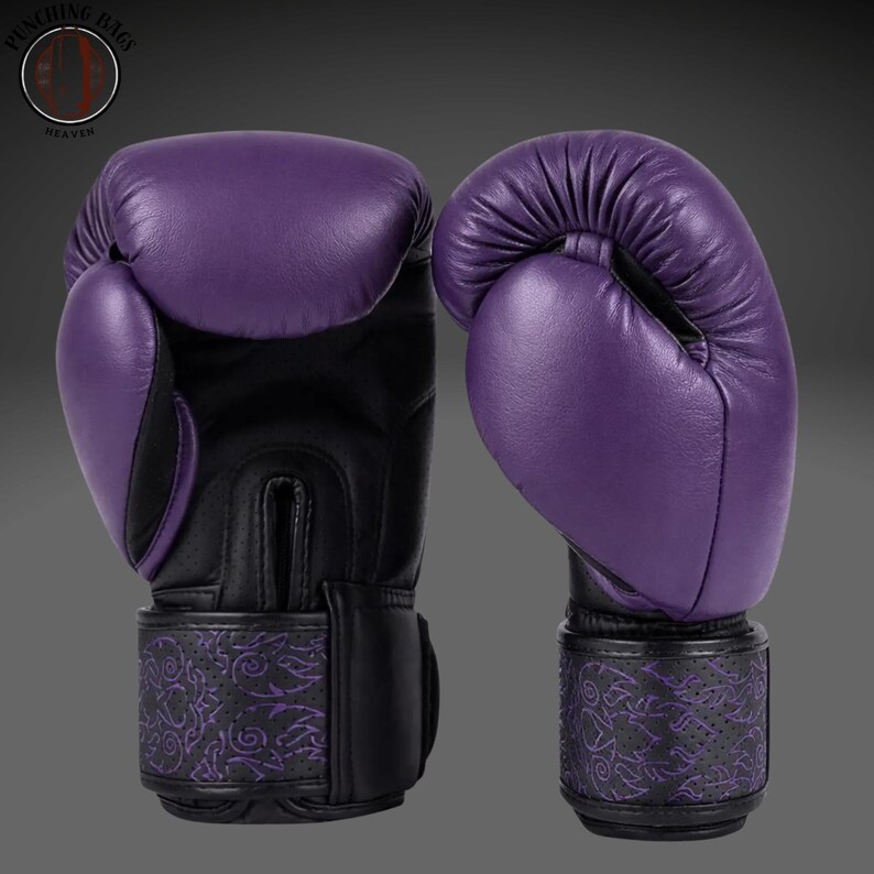 May include: A pair of purple and black boxing gloves featuring a smooth purple leather exterior on the knuckles and thumb, contrasted with black perforated leather on the palm and wrist. The wrist strap displays a detailed purple tribal pattern.