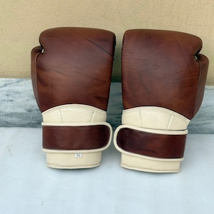 Könnte beinhalten: Ein Paar braune Leder-Boxhandschuhe mit cremefarbenen Handgelenksriemen. Die Handschuhe haben ein klassisches Design mit gepolstertem Äußeren und sicherem Handgelenkverschluss. Die Handschuhe sind für Boxen oder andere Kampfsportarten konzipiert.
