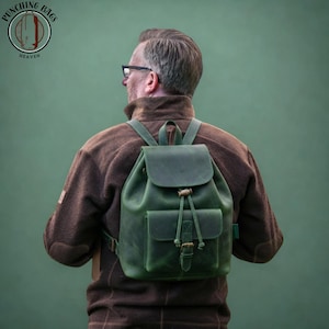 Puede incluir: Una mochila de cuero verde bosque con solapa plegable y cierre de cordón, un bolsillo frontal con correa de hebilla y correas ajustables para los hombros. El bolso tiene un acabado mate y se lleva sobre una chaqueta de forro polar marrón.