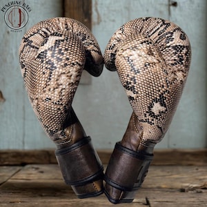 Puede incluir: Un par de guantes de boxeo con un patrón de piel de serpiente en tonos beige y negro en el cuerpo principal y un puño marrón oscuro con ribete negro. El logo en la esquina superior izquierda muestra el texto Punching Bags Heaven.