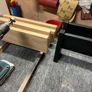 May include: A workshop scene featuring a partially constructed wooden item. The natural wood is light in colour, contrasting with a black-painted section. Various tools and materials, including sandpaper, are visible, indicating a woodworking project in progress.