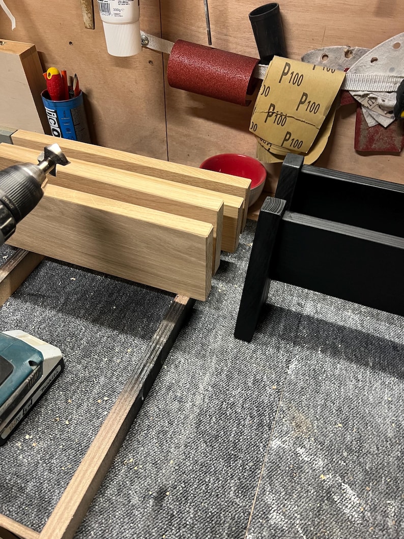 May include: A workshop scene featuring a partially constructed wooden item. The natural wood is light in colour, contrasting with a black-painted section. Various tools and materials, including sandpaper, are visible, indicating a woodworking project in progress.