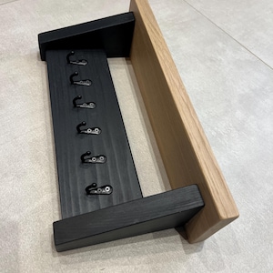 May include: A wooden key holder for wall mounting, featuring six black hooks. The design includes a black painted section for the hooks and a natural wood shelf. The style is modern and minimalist, ideal for hallways or entryways.