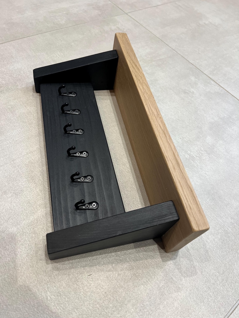 May include: A wooden key holder for wall mounting, featuring six black hooks. The design includes a black painted section for the hooks and a natural wood shelf. The style is modern and minimalist, ideal for hallways or entryways.