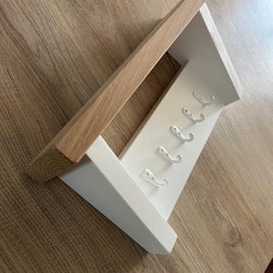May include: A wooden wall shelf with a natural wood top and a white painted base. The base has six silver hooks for hanging items. The shelf is angled and sits on a light brown wooden surface.