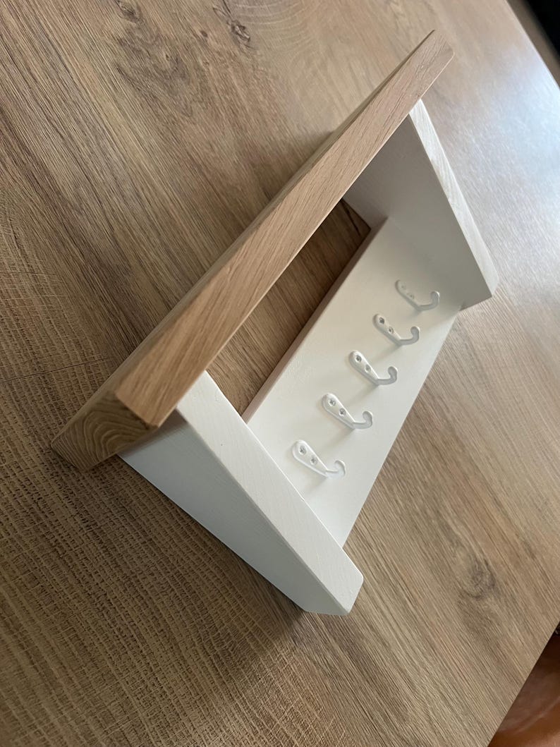May include: A wooden wall shelf with a natural wood top and a white painted base. The base has six silver hooks for hanging items. The shelf is angled and sits on a light brown wooden surface.