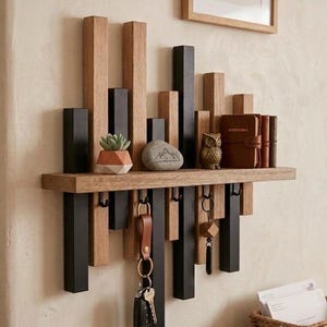 Può includere: Un portachiavi da parete in legno con un ripiano. Il design presenta elementi verticali alternati in legno nero e naturale. Il ripiano contiene una piccola succulenta, una pietra con un disegno di montagna, una statuina di gufo e un diario rilegato in pelle. Le chiavi sono appese sotto.