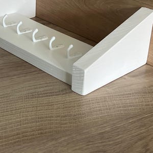 May include: A white wooden key rack featuring five white metal hooks. The rack is mounted on a light brown wooden surface. The design is simple and modern, suitable for a hallway or entryway.