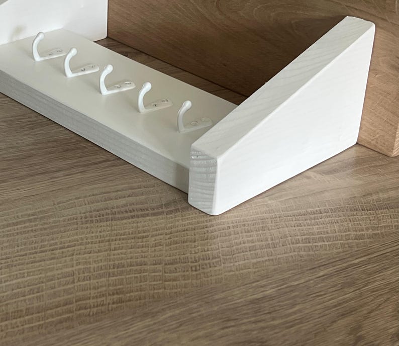 May include: A white wooden key rack featuring five white metal hooks. The rack is mounted on a light brown wooden surface. The design is simple and modern, suitable for a hallway or entryway.