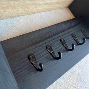 May include: A black wooden coat rack for wall mounting, featuring five black metal hooks. The rack has a rectangular design with a light-coloured wooden trim at the top. Hooks are evenly spaced along the bottom edge.