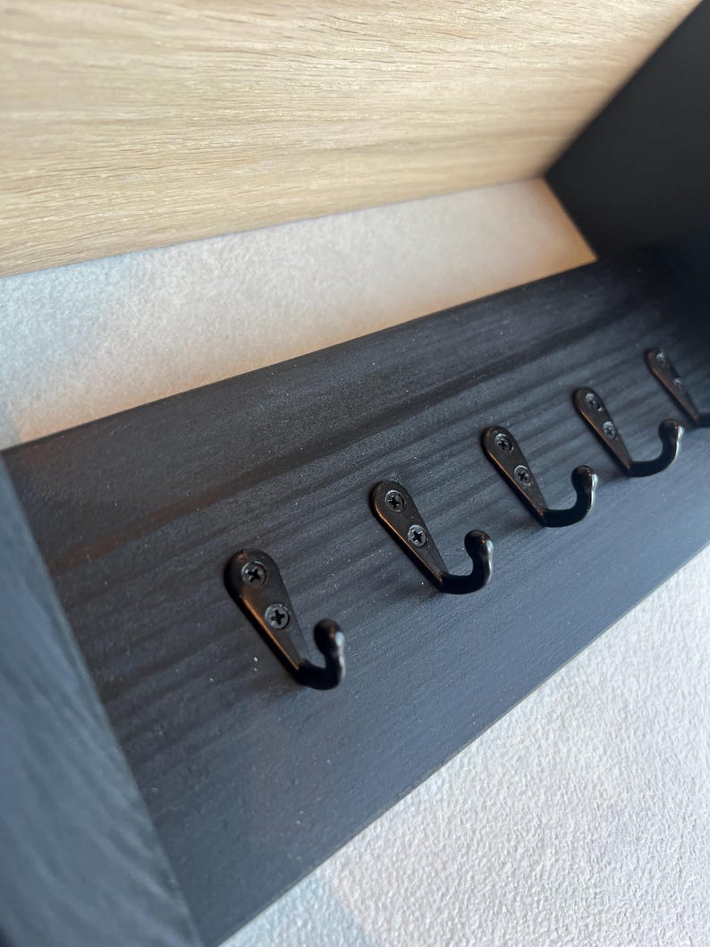 May include: A black wooden coat rack for wall mounting, featuring five black metal hooks. The rack has a rectangular design with a light-coloured wooden trim at the top. Hooks are evenly spaced along the bottom edge.