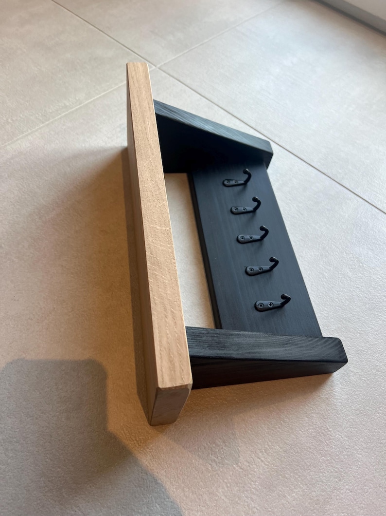 May include: A wooden key rack with a natural wood side and a black painted body. It has six metal hooks for keys. The design is simple and functional, suitable for hallway or entryway organization.