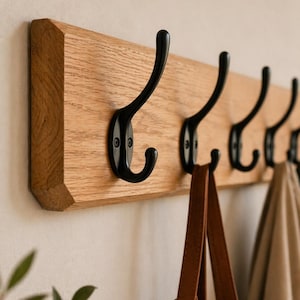 Peut inclure: Un porte-manteau en bois avec cinq crochets en métal noir. Le support est en bois de couleur claire et de forme rectangulaire. Un sac fourre-tout en cuir marron et un manteau beige sont suspendus aux crochets.