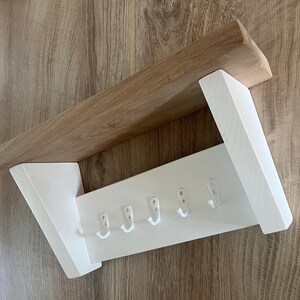 May include: A wooden wall-mounted coat rack with a natural wood shelf and white painted support and hook panel. The rack has five white hooks for hanging items. The design is simple and modern, suitable for entryways or bedrooms.
