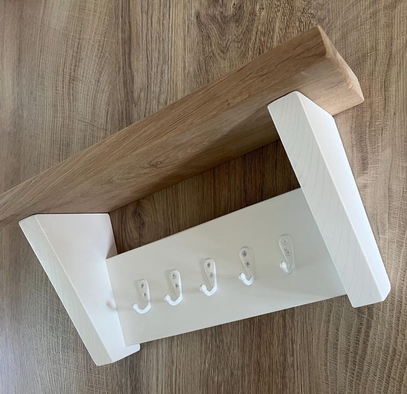 May include: A wooden wall-mounted coat rack with a natural wood shelf and white painted support and hook panel. The rack has five white hooks for hanging items. The design is simple and modern, suitable for entryways or bedrooms.