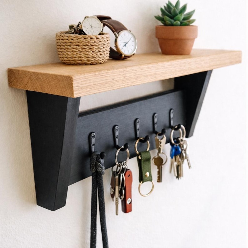 May include: A wooden wall shelf with a black key rack. The shelf has a natural wood finish and holds a small potted plant, a woven basket, and two watches. Keys hang from the hooks below.