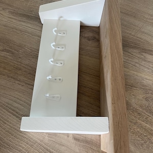 May include: A white wooden wall-mounted coat rack with six hooks. The rack has a shelf and a sloped top. The right side is made of natural wood. The hooks are white plastic. The rack is designed for hanging coats, hats, or other items.