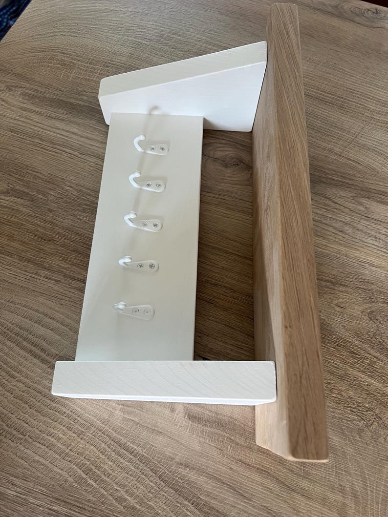 May include: A white wooden wall-mounted coat rack with six hooks. The rack has a shelf and a sloped top. The right side is made of natural wood. The hooks are white plastic. The rack is designed for hanging coats, hats, or other items.