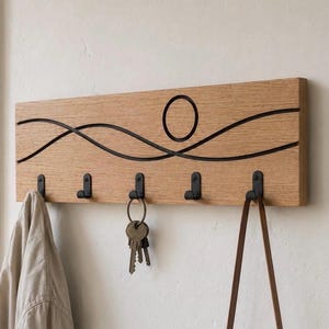Può includere: Un appendiabiti da parete rettangolare in legno chiaro con cinque ganci in metallo nero. La superficie in legno è decorata con due linee ondulate nere orizzontali che si incrociano e un cerchio nero centrale. Un mazzo di chiavi pende dal gancio centrale.
