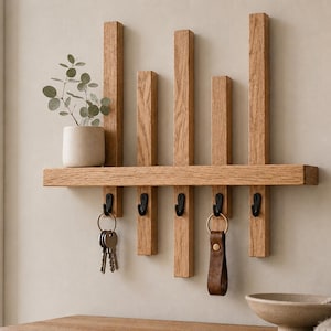 May include: A wall-mounted light brown wooden shelf featuring five vertical slats of varying heights and five black metal hooks. A small light-coloured ceramic pot with a green plant sits on the left side of the shelf, while keys and a leather fob hang from the hooks.